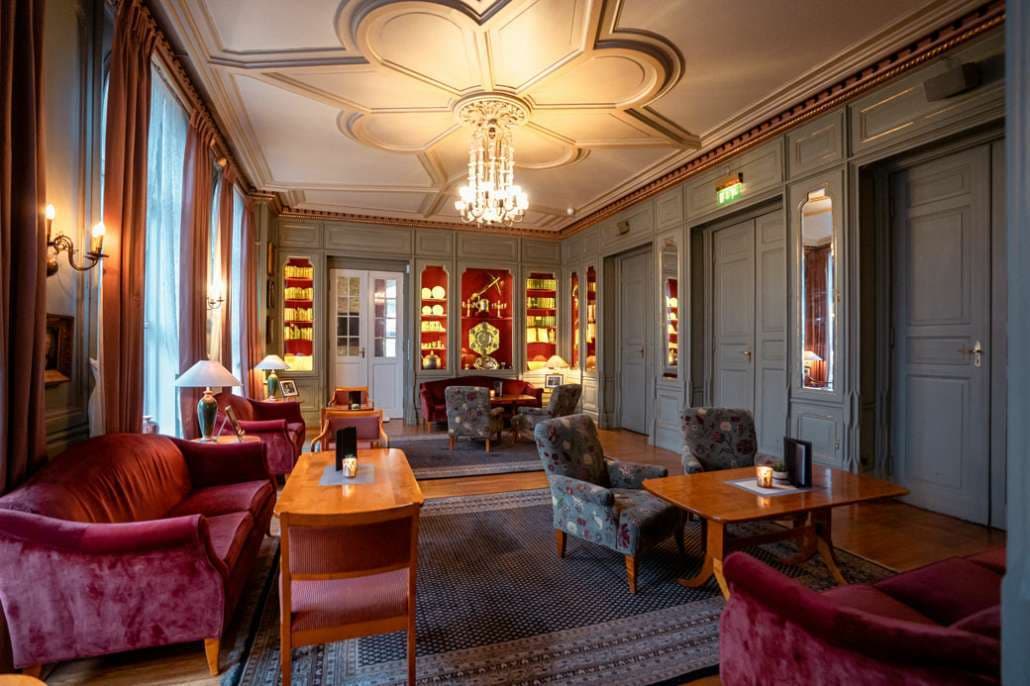 Salong med grågrønne vegger, sofaer, stoler, og lysekrone.