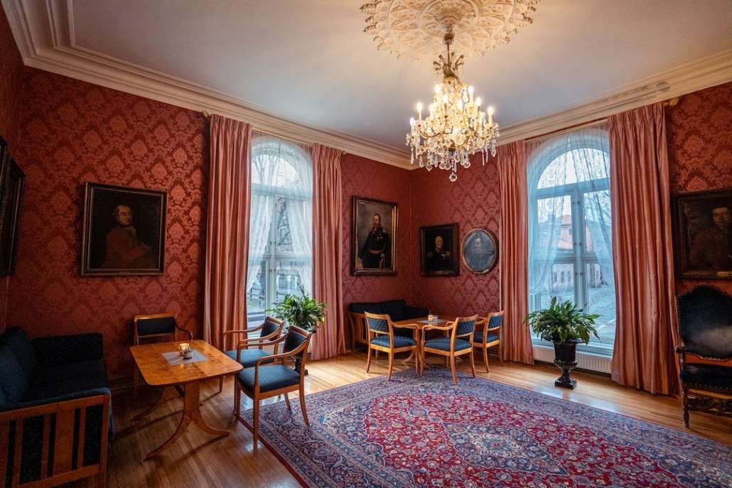Elegant salong med rød tapet, lysekrone og flotte møbler i tre med blått trekk.